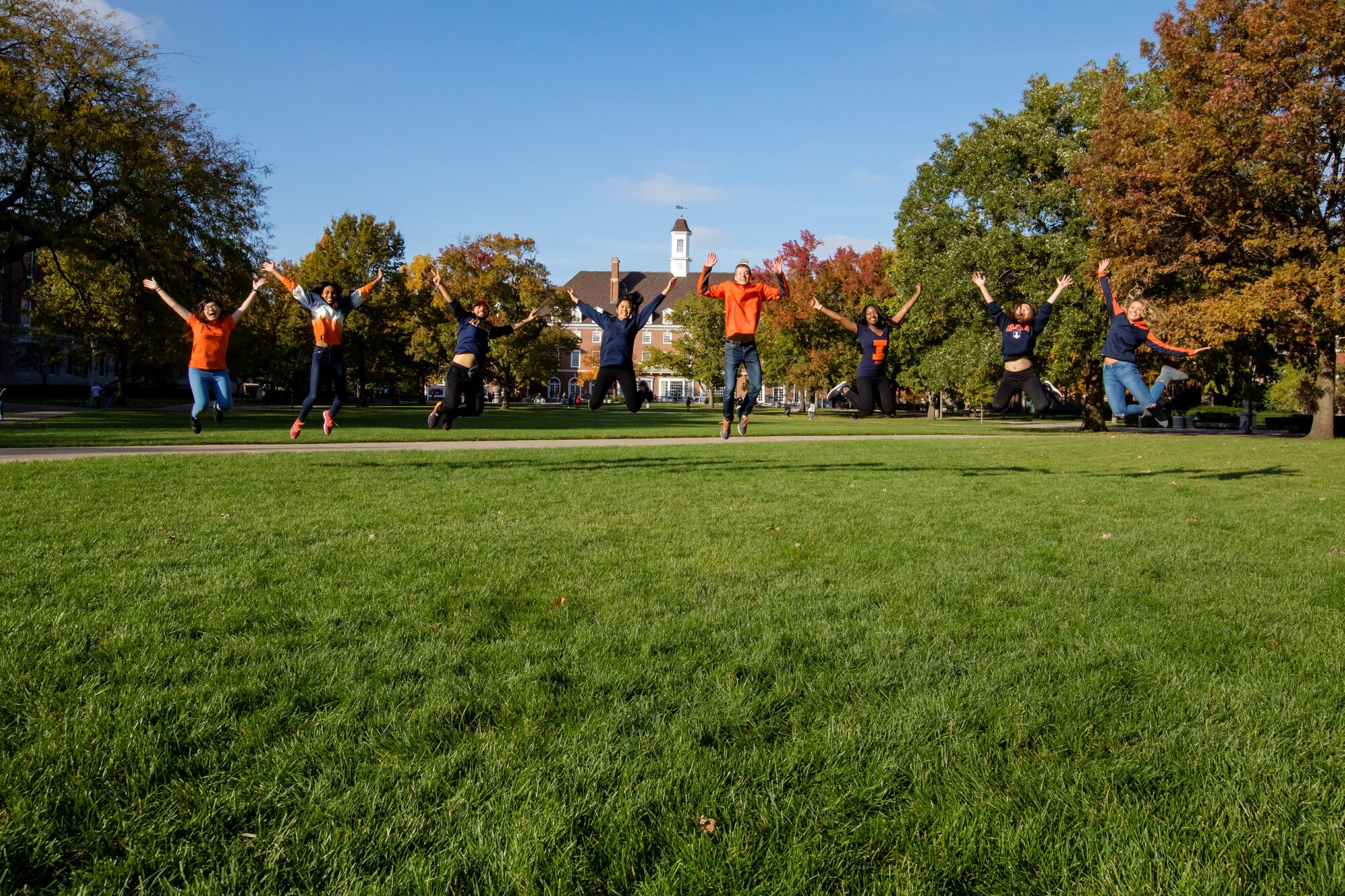 Diverse group of students wearing Illini gear jumping in the air on the quad in front of the Illini Union building, with trees beginning to turn to Fall colors